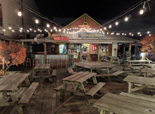 A rustic outdoor seating area with wooden tables and string lights, featuring a bar named "Barnacle Buds."