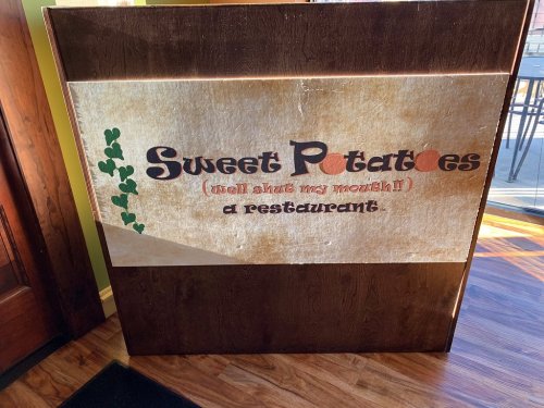 Sign for "Sweet Potatoes" restaurant with a playful tagline, displayed on a wooden background.