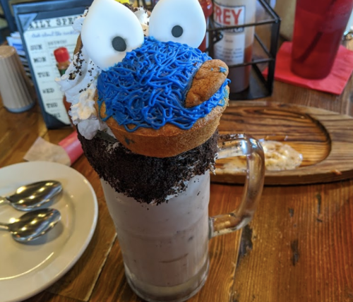 A large milkshake topped with a cookie monster donut, whipped cream, and crushed cookies in a glass mug.