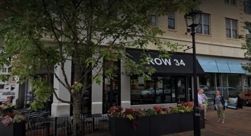A restaurant with outdoor seating, surrounded by trees and flowers, on a busy street with pedestrians.