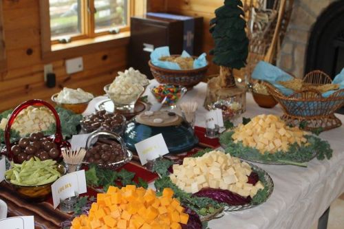 A festive table spread with various cheeses, snacks, and candies, decorated with greenery and a small tree centerpiece.