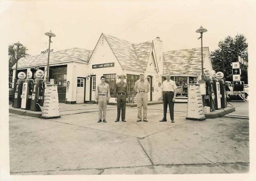 Historic black-and-white photo of four men standing in front of a vintage gas station with old fuel pumps.