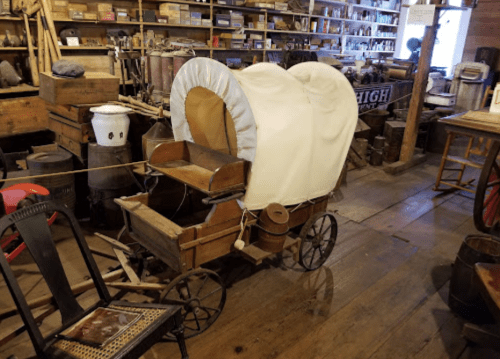 A vintage wooden child's wagon with a white canopy, displayed in a rustic shop filled with various antique items.