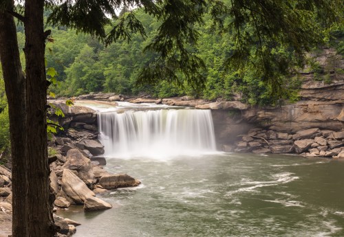 A serene waterfall cascades over rocks into a calm river, surrounded by lush green trees and foliage.