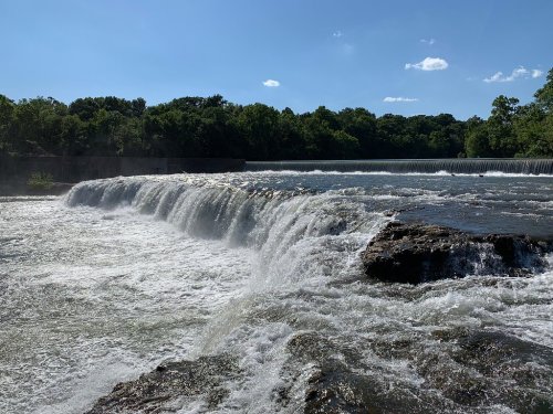 A waterfall cascades over rocks under a clear blue sky, surrounded by lush green trees.