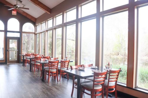 Bright restaurant interior with wooden tables and chairs, large windows overlooking a serene outdoor view.
