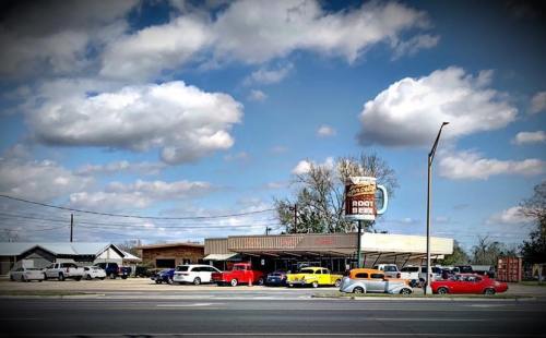 A retro-style root beer stand with colorful cars parked outside, under a blue sky with fluffy clouds.