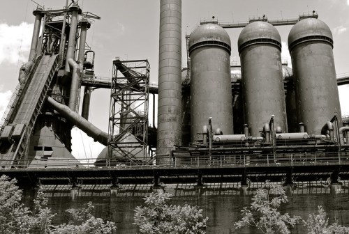 Black and white image of an industrial facility with large silos and pipes against a cloudy sky.