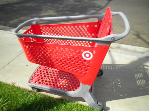 A red shopping cart with a perforated design parked on a sidewalk next to grass.