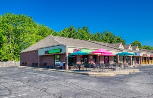 A brick storefront with colorful signs for frozen yogurt, featuring outdoor seating and umbrellas on a sunny day.