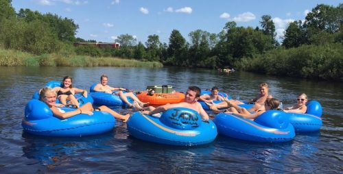 A group of people relaxes on blue inner tubes in a river on a sunny day, surrounded by trees and a clear sky.