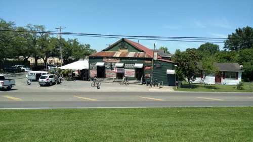 A rustic building with a green exterior and red roof, surrounded by trees and parked cars on a sunny day.