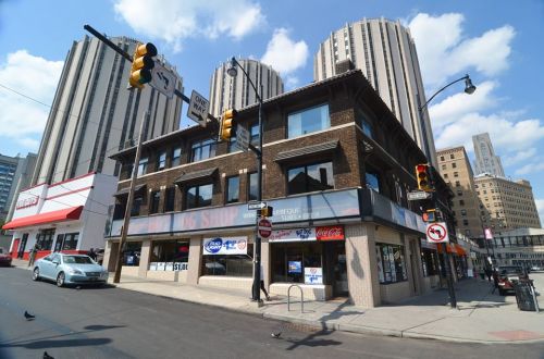 A brick building with large windows and signage, located at a busy intersection with tall buildings in the background.