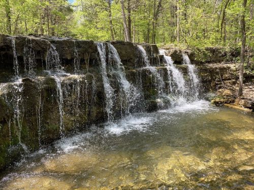 A serene waterfall cascading over rocky ledges into a clear pool, surrounded by lush green trees.