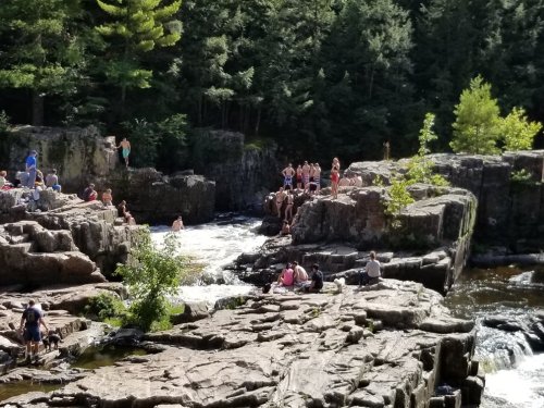 A group of people enjoying a sunny day by a rocky river with cascading water, surrounded by trees.