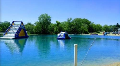 A sunny lake scene featuring inflatable water slides and obstacles, surrounded by trees and a sandy beach.