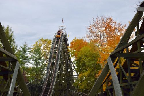 A roller coaster climbs a steep track surrounded by colorful autumn trees under a cloudy sky.