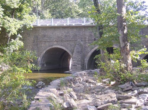 A stone bridge with arches over a calm stream, surrounded by trees and rocky banks.