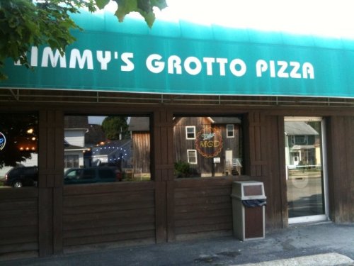 Sign for "Jimmy's Grotto Pizza" on a green awning above a wooden storefront with a trash can nearby.