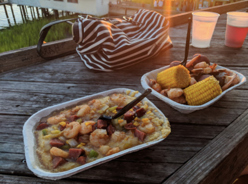 A seafood dish with shrimp, sausage, and corn, alongside a striped bag and drinks on a wooden table at sunset.