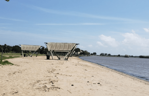 Three shaded structures on a sandy beach by a calm river under a clear blue sky.
