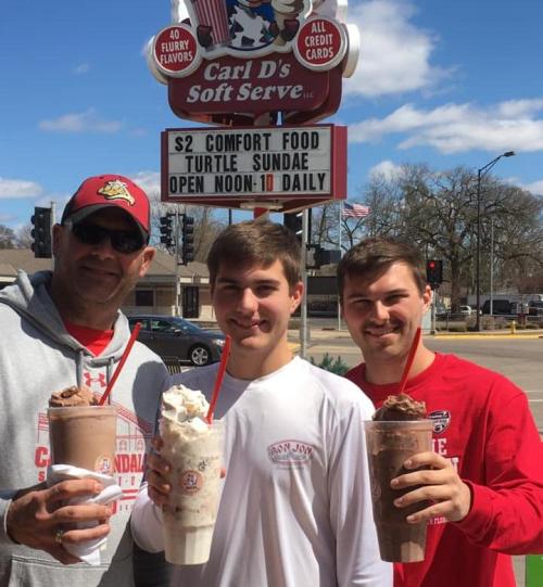 Three people hold large milkshakes in front of a sign for Carl's Soft Serve, promoting $2 comfort food and turtle sundaes.