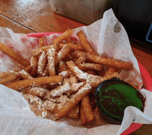 A basket of golden-brown fries dusted with powdered sugar, served with a small cup of green dipping sauce.