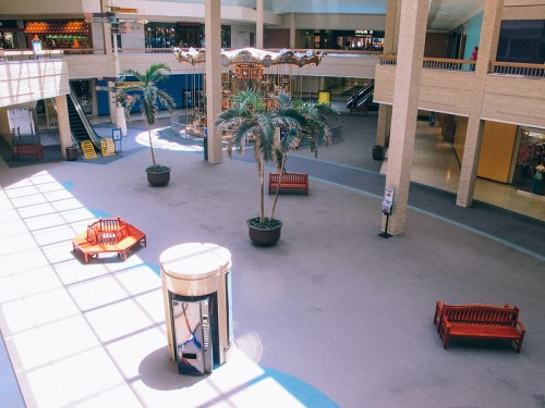 A spacious indoor area with palm trees, red benches, and a carousel in the background, illuminated by natural light.