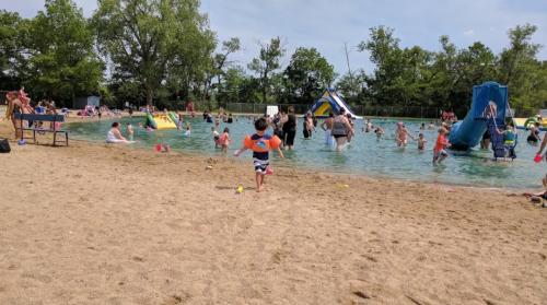 A sandy beach area with people swimming and playing in a lake, including a child running with an orange float.