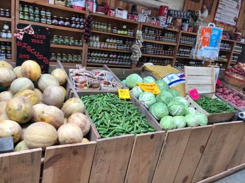 A vibrant display of fresh vegetables and melons in a market, with jars of preserves in the background.