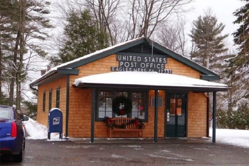 A small, wooden post office building with a snow-covered roof, surrounded by trees in a winter setting.