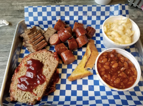 A tray with sliced meatloaf topped with sauce, sausage pieces, macaroni and cheese, baked beans, and a slice of bread.