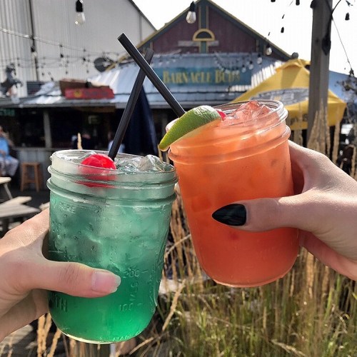 Two hands hold colorful cocktails in mason jars, one blue with a cherry and the other orange with a lime slice.
