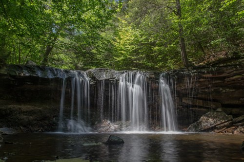 A serene waterfall cascades over rocks, surrounded by lush green trees and a calm pool of water below.
