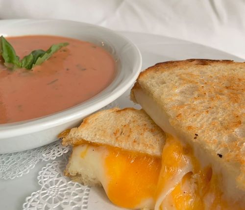 A close-up of a grilled cheese sandwich with melted cheese beside a bowl of tomato soup garnished with basil.