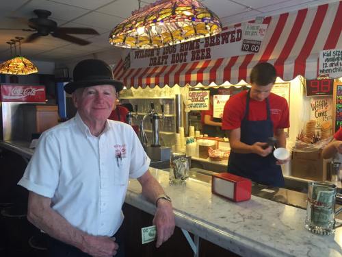 An older man in a bowler hat stands at a diner counter, while a young employee serves customers in the background.