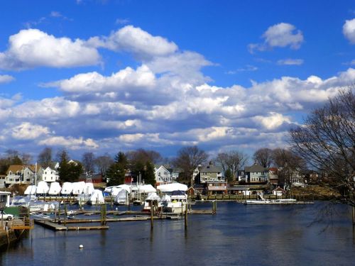 A serene waterfront scene with boats docked, white tents, and charming houses under a blue sky with fluffy clouds.