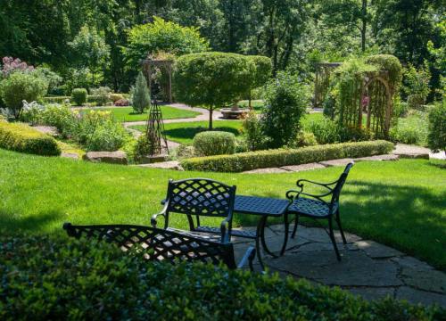 A serene garden with lush greenery, stone paths, and decorative seating areas surrounded by vibrant plants.