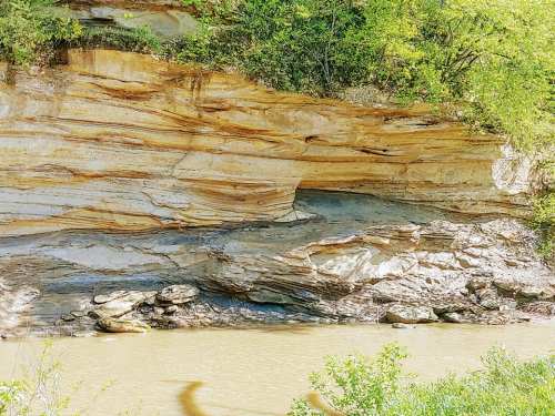 Layered rock formations beside a calm river, surrounded by greenery and trees.