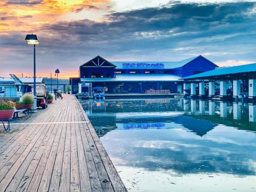 A serene waterfront scene at sunset, featuring a wooden dock, calm water, and a blue building reflecting in the lake.