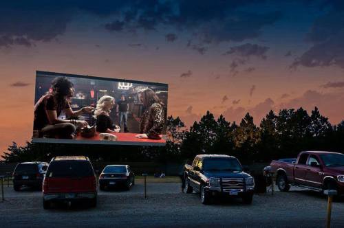 A drive-in movie theater at sunset, featuring a film on a large screen with parked cars in the foreground.