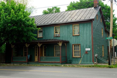 A two-story green wooden house with a porch, located at the corner of a street, surrounded by trees.