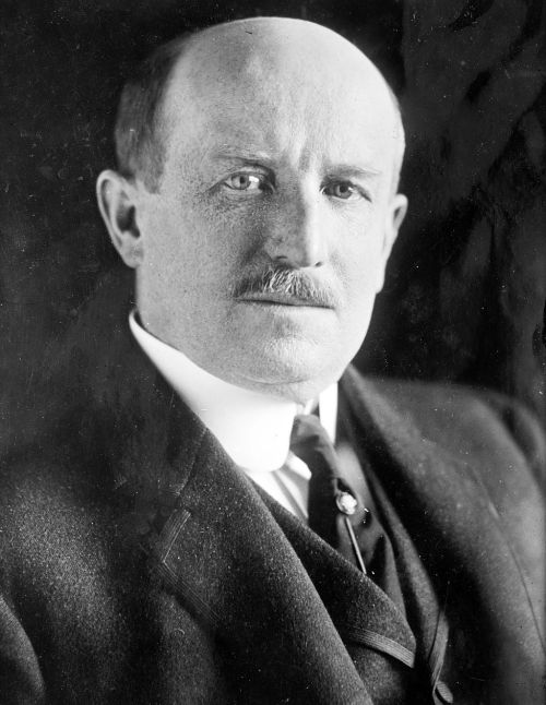 A black-and-white portrait of a man with a mustache, wearing a suit and tie, looking directly at the camera.