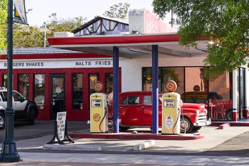 A vintage diner with a red car and classic gas pumps, featuring a sign for burgers, shakes, and fries.