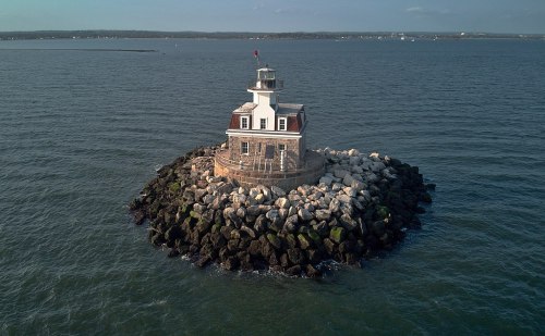 A lighthouse stands on a rocky islet surrounded by calm waters, with a clear sky in the background.