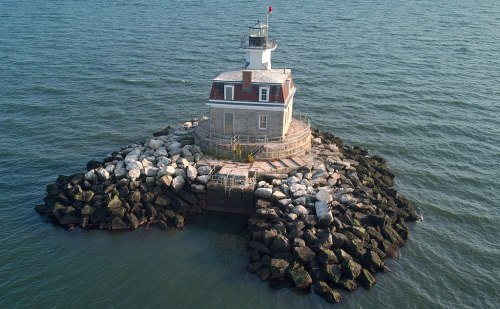 A small lighthouse on a rocky island surrounded by water, with a red flag flying atop its roof.