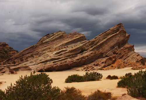 A dramatic rock formation rises against a cloudy sky, surrounded by sparse vegetation in a desert landscape.