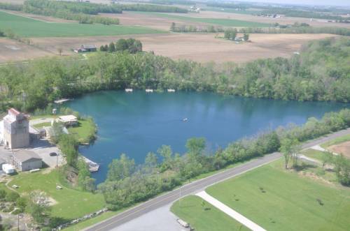 Aerial view of a serene blue lake surrounded by greenery and farmland, with a small building and a road nearby.