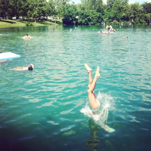 A person performs a handstand dive into a clear lake, surrounded by others swimming and enjoying the water.