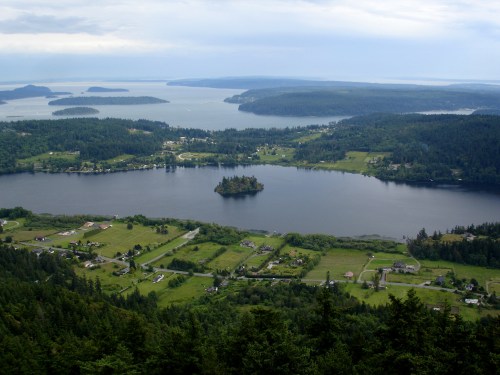 Aerial view of a lush green landscape with a lake, islands, and distant hills under a cloudy sky.
